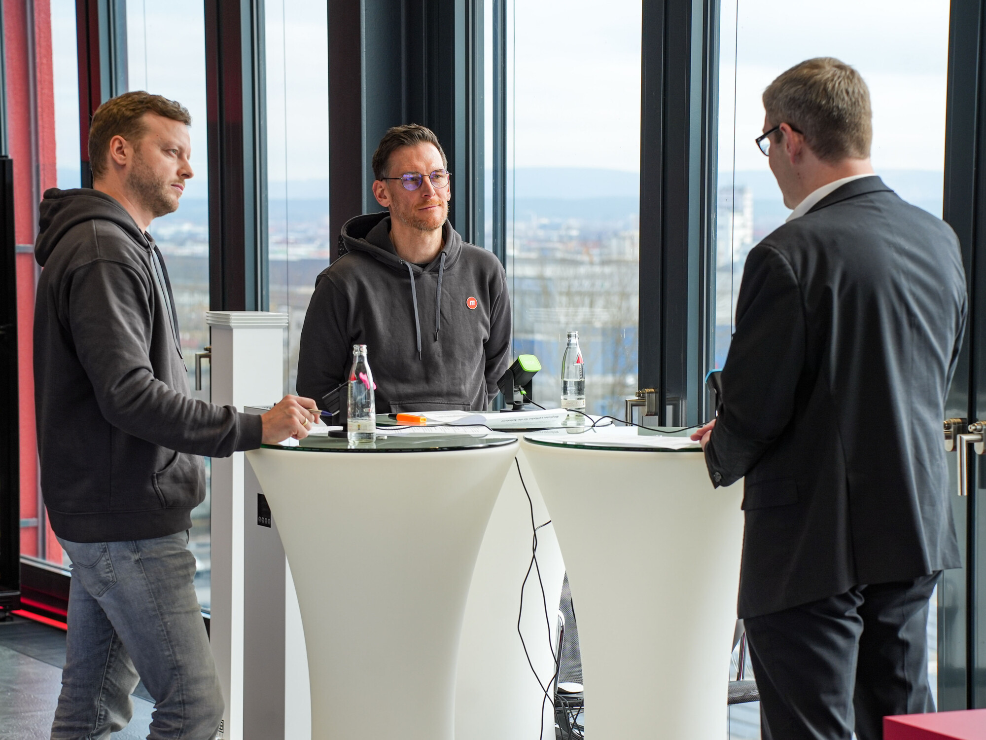 Drei Männer im Gespräch, zwei leger in Hoodies, einer im Anzug, an Stehtischen mit Stadtblick und Wasserflaschen.