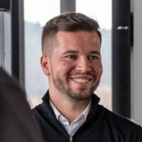 Junger Mann mit Bart und lächelndem Gesichtsausdruck in Büro mit natürlichem Licht und moderner Ausstattung.