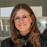 Frau mit langen braunen Haaren, moderner Brille und schwarzem Outfit lächelt freundlich in hellem Innenraum.