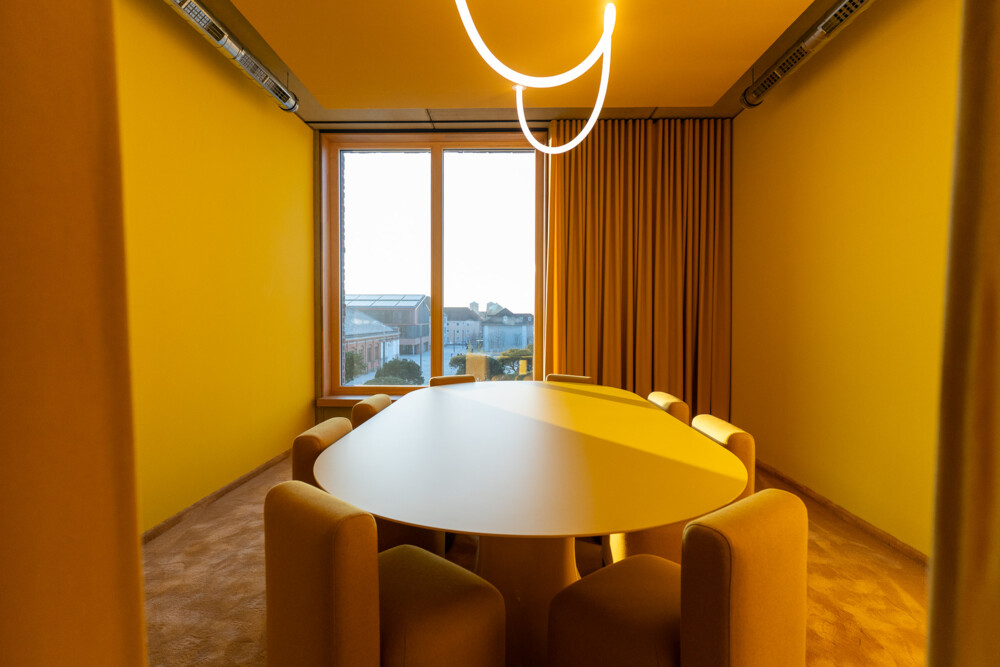 Moderner, gelb gestalteter Meetingraum mit ovalem Konferenztisch, gepolsterten Stühlen und großem Fenster mit Blick auf Stadt.