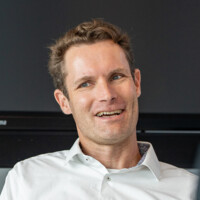 Mann mit weißem Hemd lächelt entspannt vor dunklem Hintergrund, freundlich und kommunikativ wirkend.