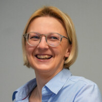 Frau mit blonden Haaren und Brille lächelt freundlich in hellblauem Hemd vor neutralem Hintergrund.