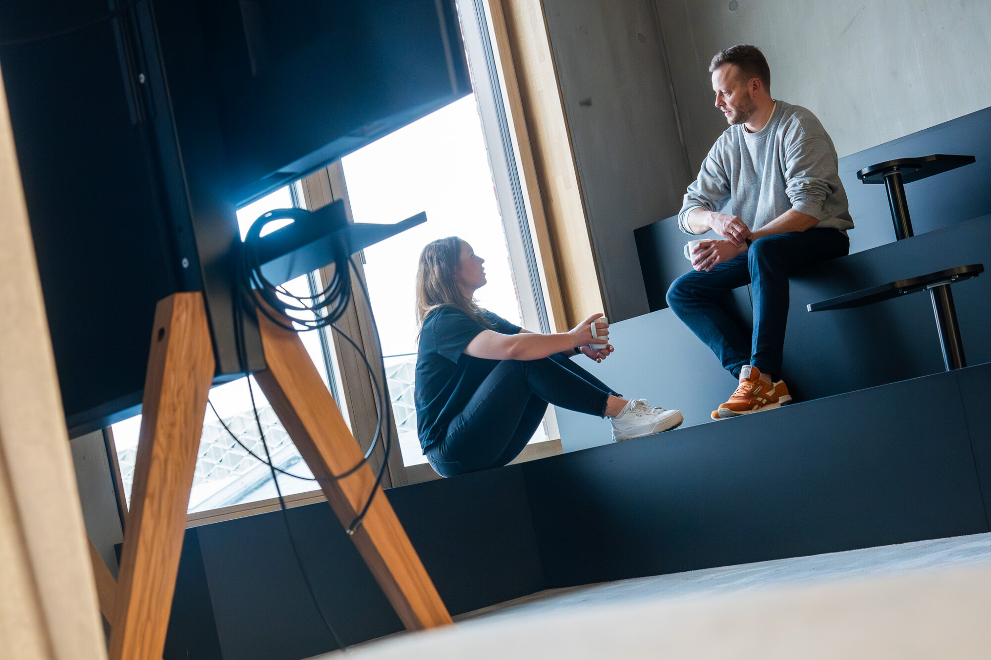 Zwei junge Menschen im modernen Büro unterhalten sich entspannt auf einer Sitzbank am Fenster mit Tageslicht.