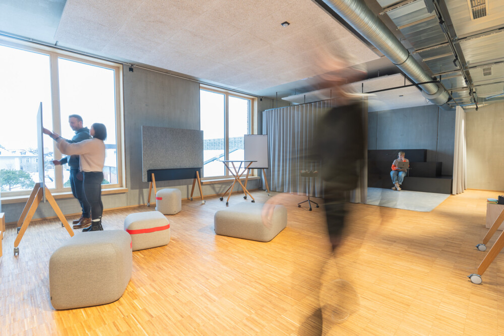 Moderne Bürofläche mit großen Fenstern, kreativen Arbeitsbereichen und Mitarbeitern beim Brainstorming und Lesen.