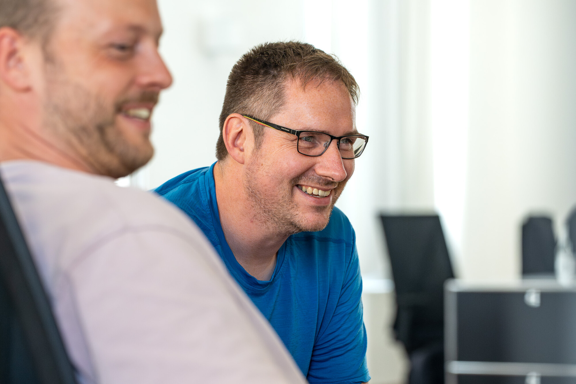 Zwei Männer im Büro im Gespräch, einer trägt blaue Shirt und Brille, entspannte und freundliche Atmosphäre.