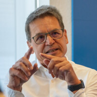 Mann mit grauen Haaren und Brille erklärt etwas mit beiden Händen, trägt weißes Hemd und schwarzen Fitness-Tracker.