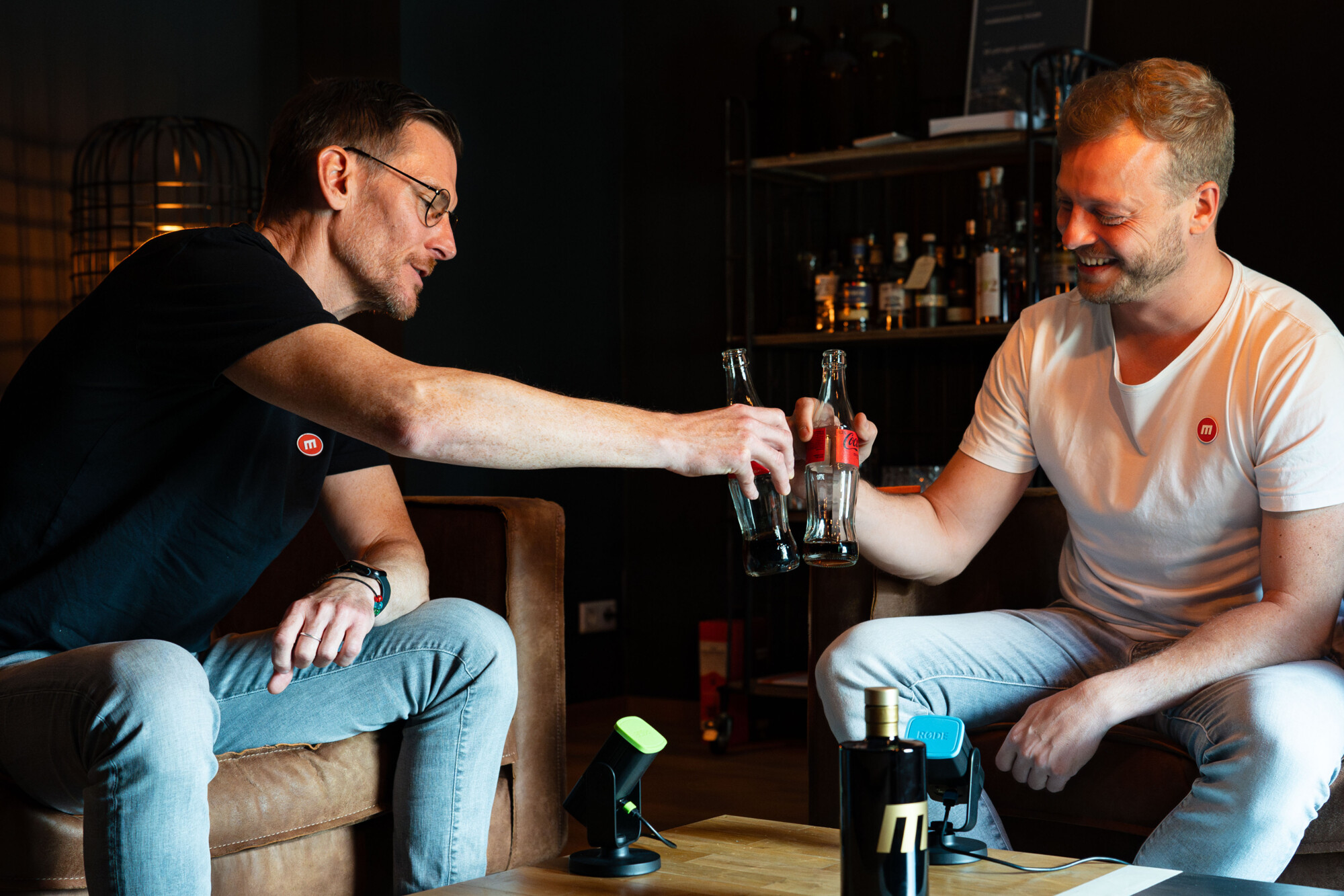 Zwei Männer in entspannter Atmosphäre stoßen mit Glasflaschen an, lachend und gemütlich in modernem Raum.