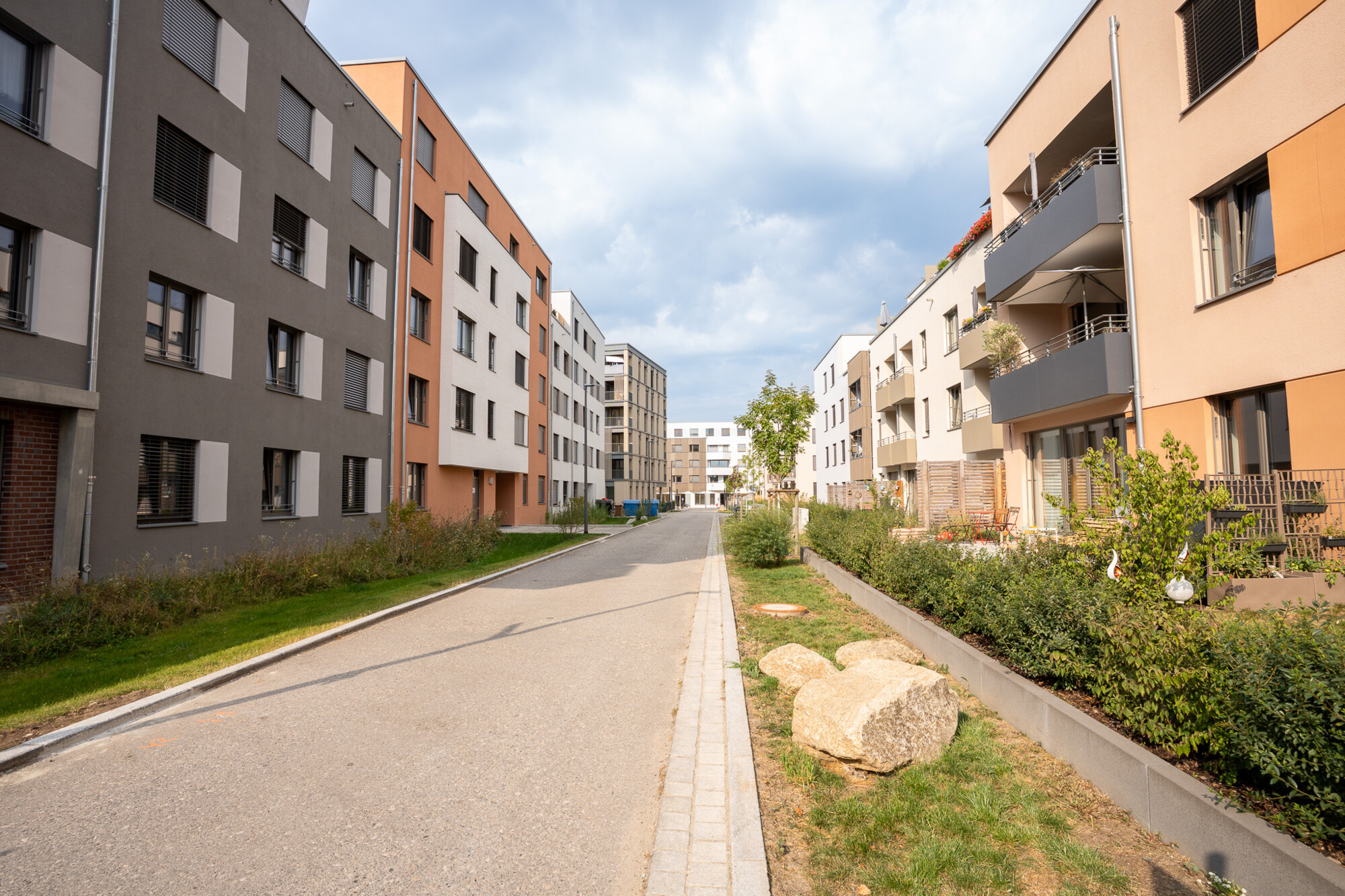 Moderne Mehrfamilienhäuser mit Balkonen und Grünanlagen entlang ruhiger Straße unter bewölktem Himmel in Neubaugebiet.
