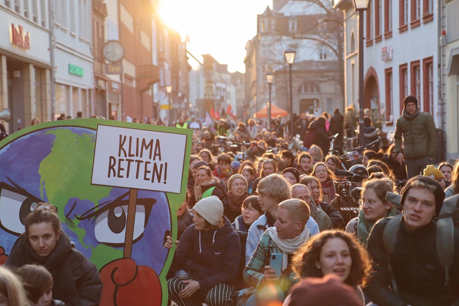 Menschenmenge bei sonniger Klimademonstration mit Schild „Klima Retten!“ und großer Umwelt-Erde-Grafik auf belebter Straße.
