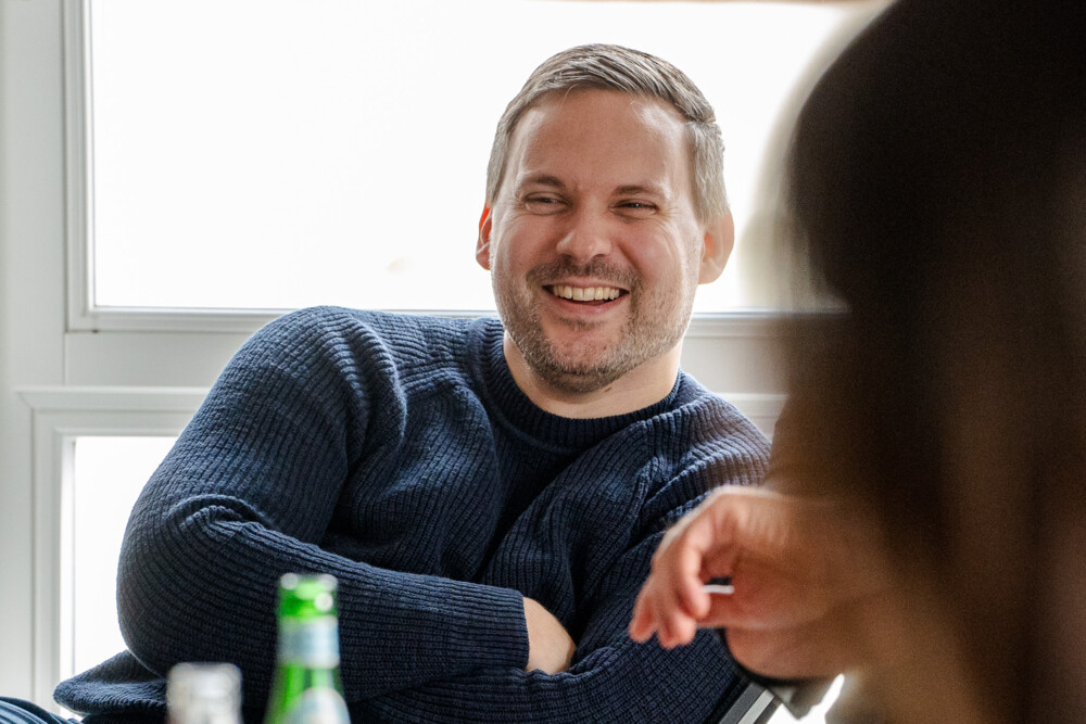 Mann in blauem Pullover lacht entspannt bei zwanglosem Gespräch in heller Büroatmosphäre.