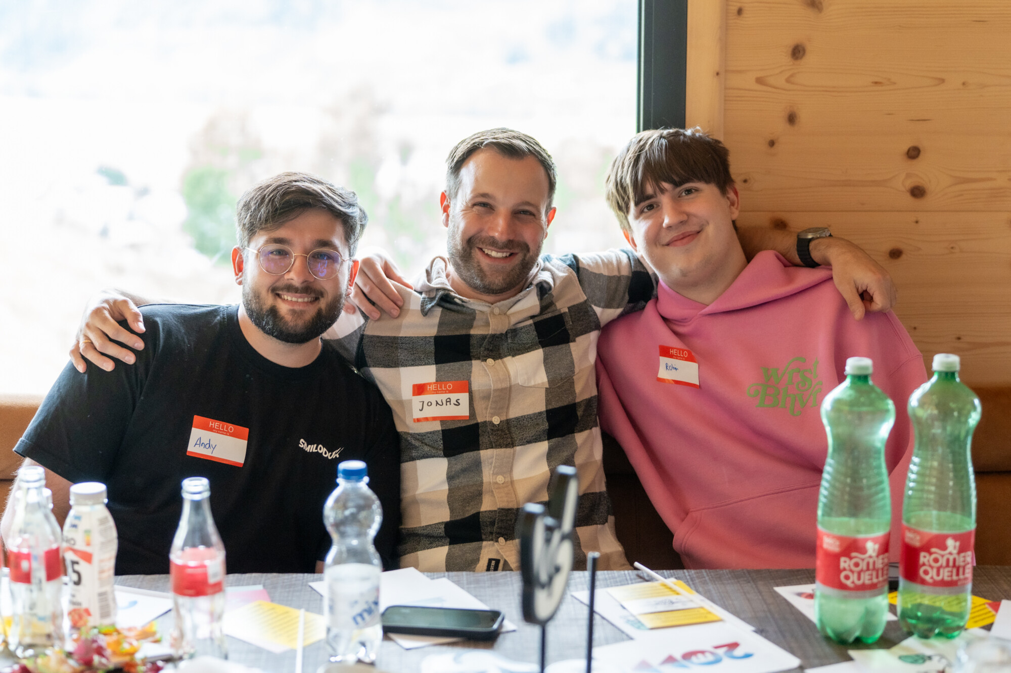 Drei junge Männer mit Namensschildern lächeln beim gemeinsamen Workshop an einem Tisch mit Getränken und Unterlagen.