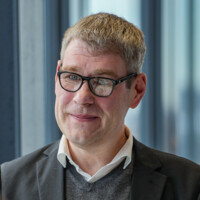 Mann mit Brille und grauem Sakko lächelt freundlich vor hellem, modernem Bürofenster.