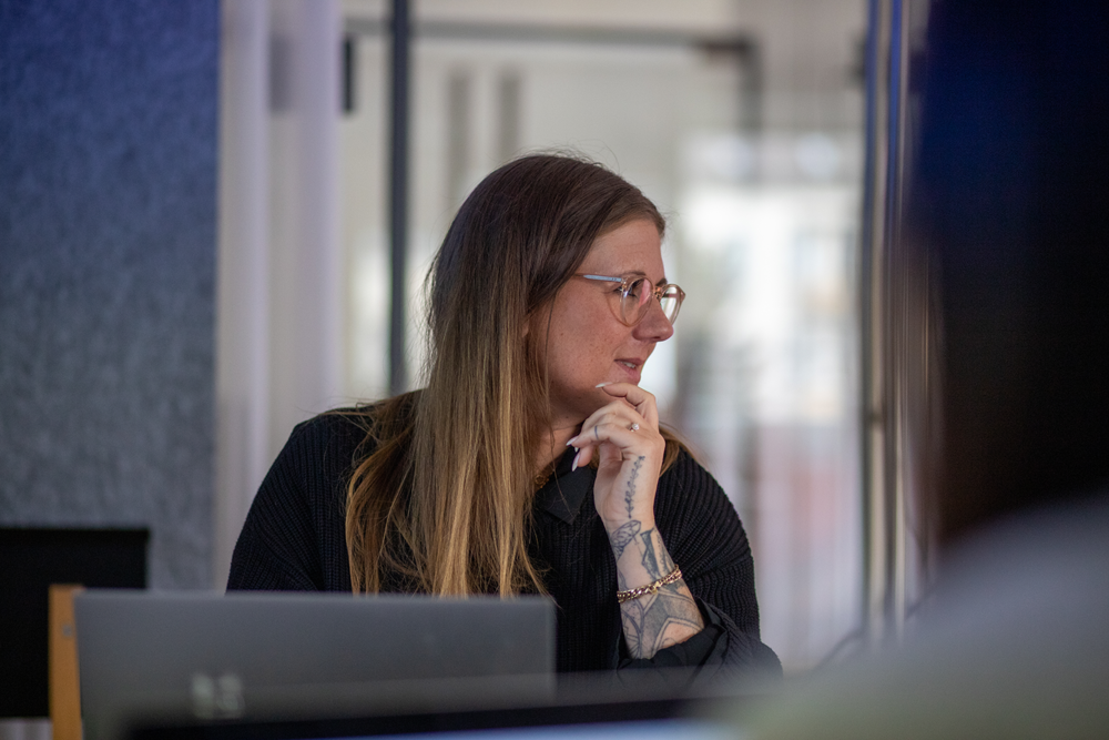 Frau mit Brille und Tattoos sitzt nachdenklich am Laptop in hellem Büro und schaut seitlich vorbei.