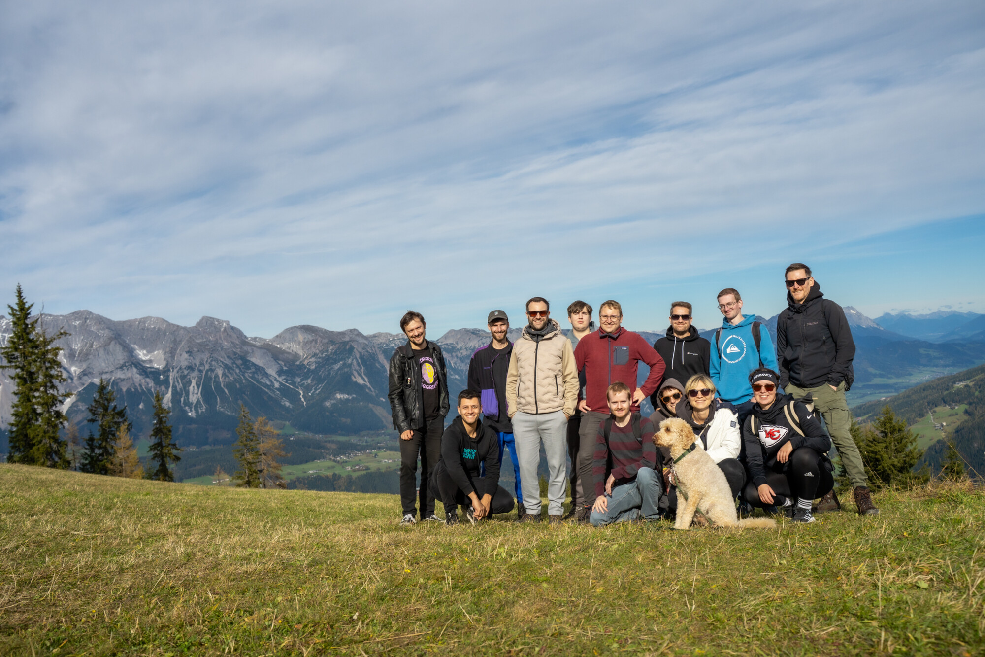 Kollegengruppe mit Hund bei Wanderung vor Bergkulisse in sonniger Alpenlandschaft, Freizeit und Teambuilding