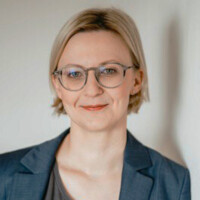 Frau mit blonden kurzen Haaren und Brille, professionell und freundlich lächelnd vor hellem Hintergrund