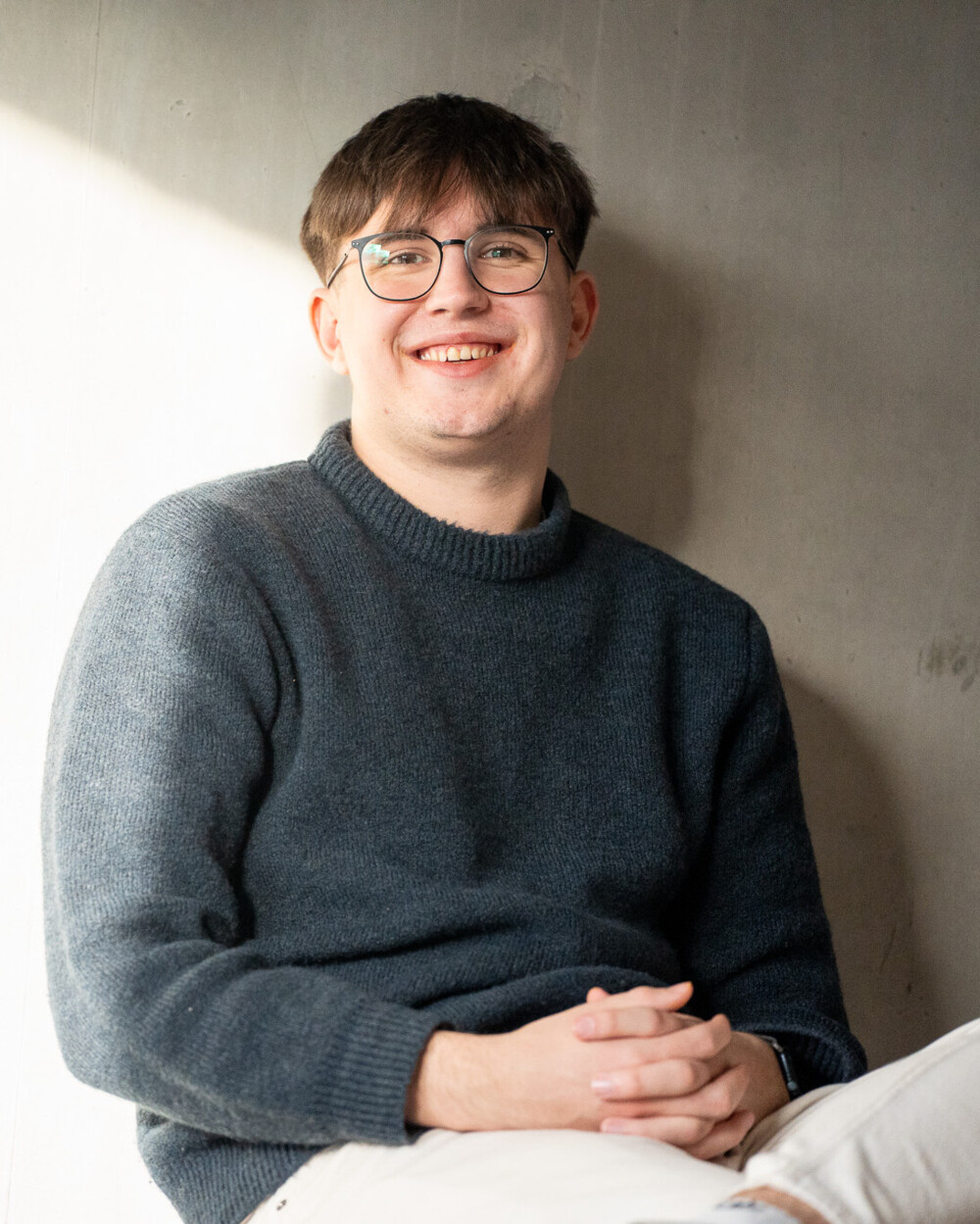 Junger Mann mit Brille und grauem Pullover sitzt lächelnd vor grauer Wand, entspannte Atmosphäre.