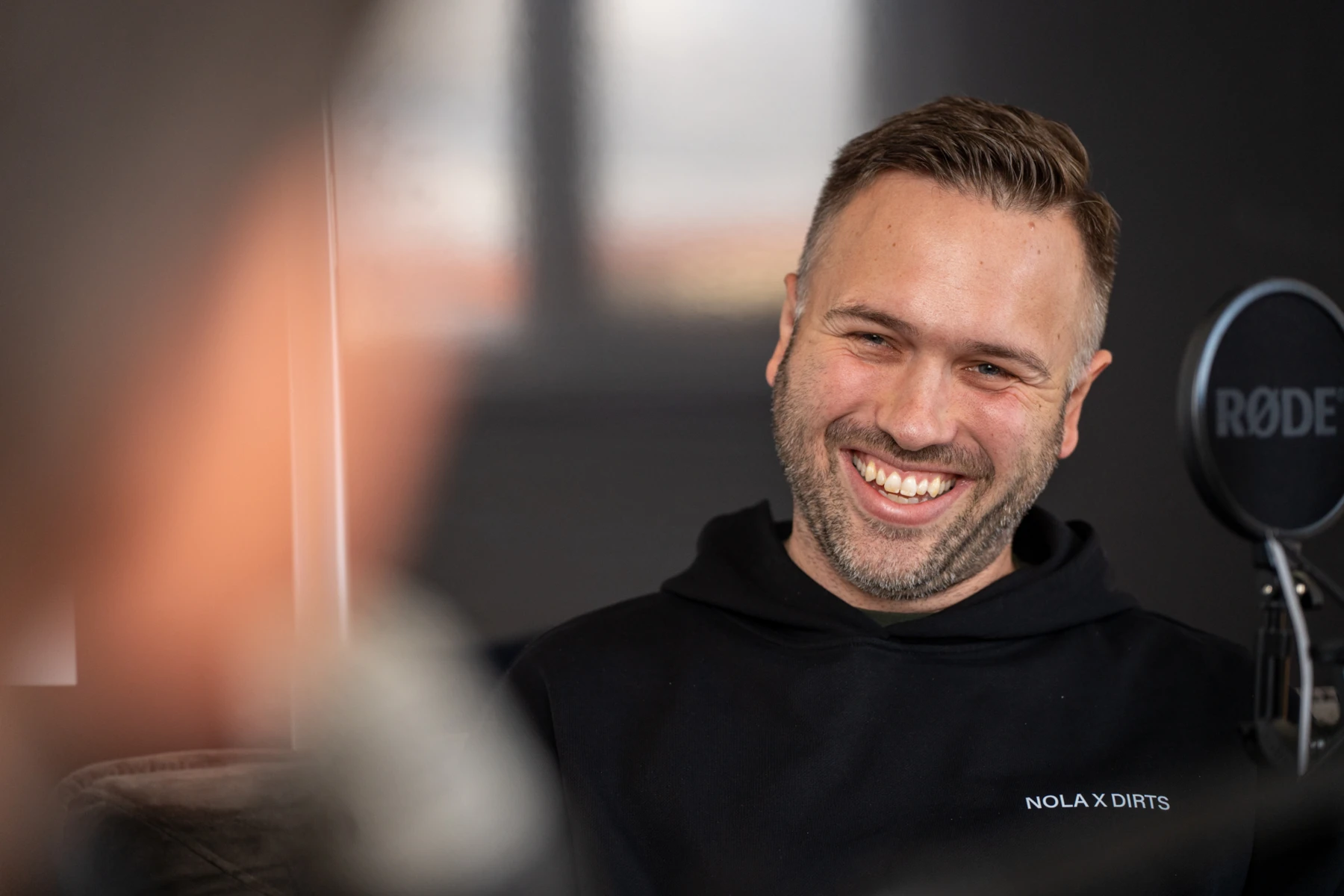 Mann mit Kapuzenpullover lacht entspannt vor Mikrofon in moderner Podcast-Atmosphäre.