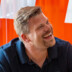 Mann mit blondem Haar und Bart lacht herzlich vor orange-weißem Hintergrund, vermittelt positive Stimmung und Offenheit.