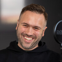 Mann mit kurzem braunem Haar und Bart lacht vor Mikrofon in modernem Studio, freundliche Atmosphäre