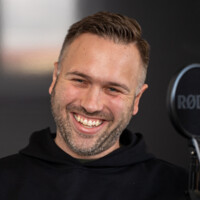Lachender Mann mit kurzem Haar und Bart vor Mikrofon, entspanntes Podcast- oder Videostreaming-Setup