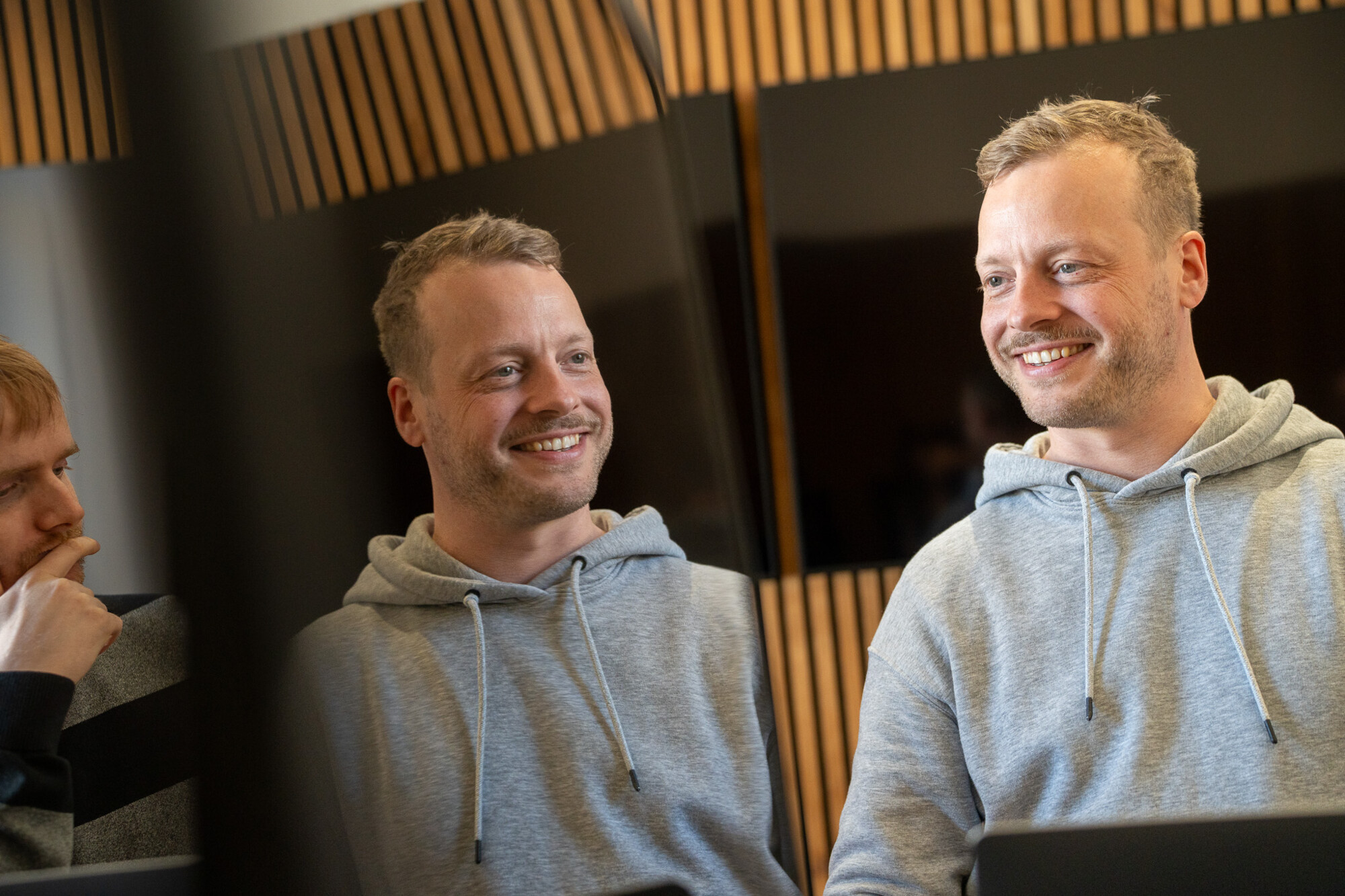 Zwei Männer im Gespräch, einer trägt grauen Kapuzenpullover, entspannte Atmosphäre in modernem Büro mit Holzverkleidung.