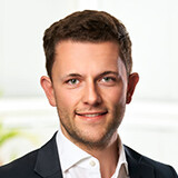 Junger Mann mit braunen Haaren und blauem Hemd lächelt freundlich vor hellem Hintergrund