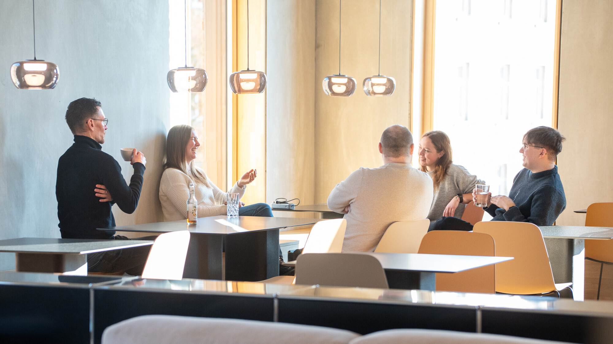 Fünf Personen in hellem, modernem Büro lachen und unterhalten sich entspannt bei Getränken am Tisch.