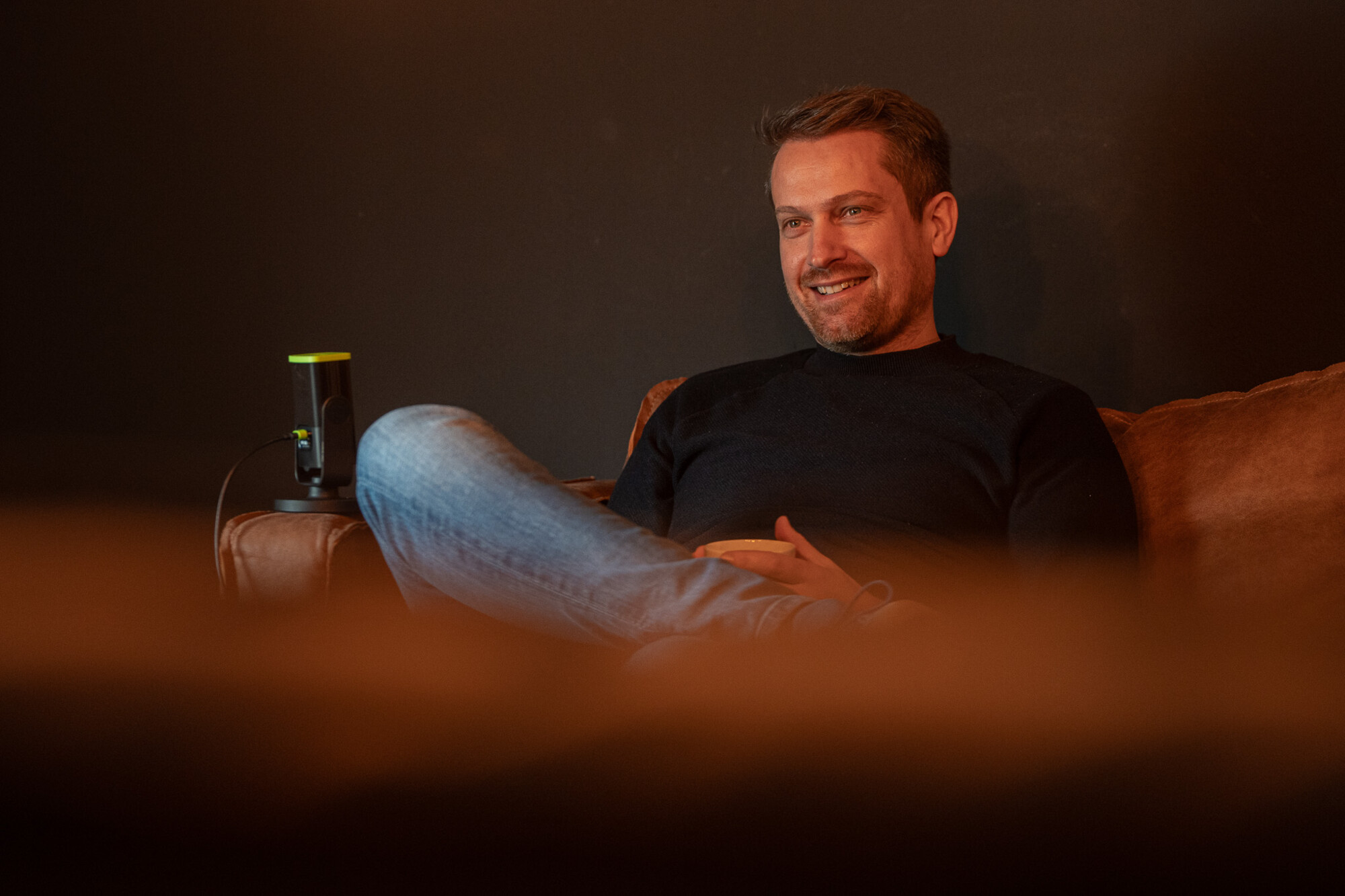 Mann entspannt lächelnd auf braunem Sofa vor dunkler Wand, locker gekleidet mit Jeans und schwarzem Pullover.