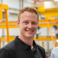 Lächelnder Mann mit rotem Haar in schwarzem Poloshirt in industrieller Produktionshalle, modernes Arbeitsumfeld.