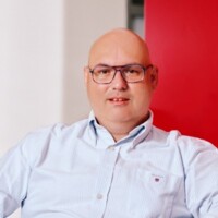 Mann mit Brille und hellblauem Hemd vor rotem Hintergrund, freundlich und professionell wirkend.