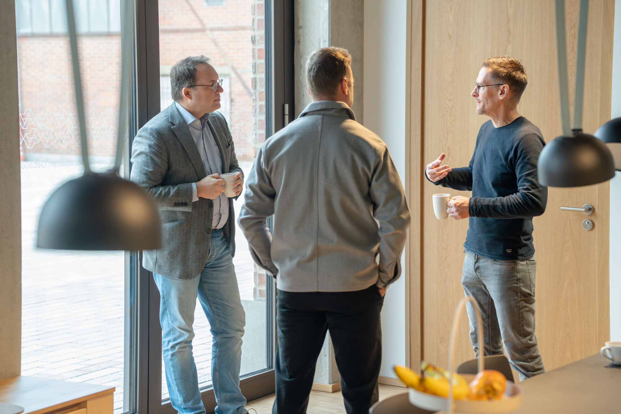 Drei Männer im Gespräch bei Kaffeepause in modernem Büro mit Tageslicht und entspanntem Arbeitsklima.