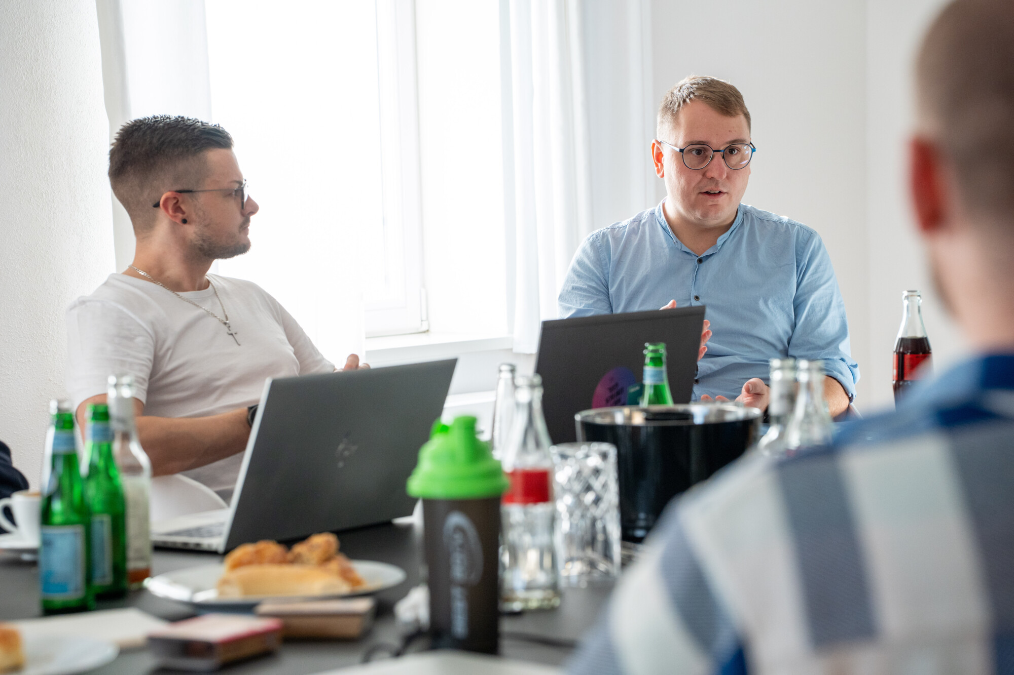 Drei Männer in Besprechung mit Laptops und Getränken, konzentrierter Austausch in hellem Büro.