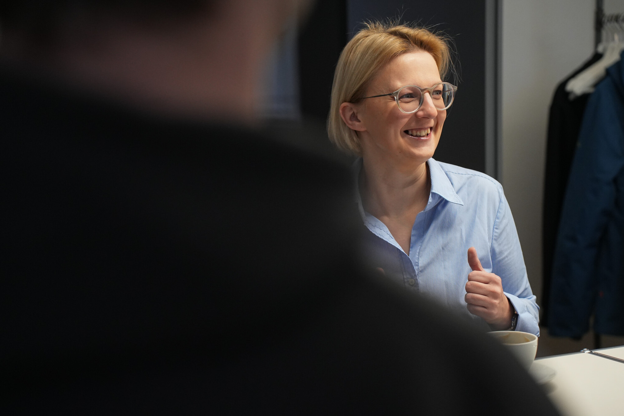 Frau mit Brille und blauem Hemd lächelt und gibt Daumen hoch in moderner Büroumgebung.