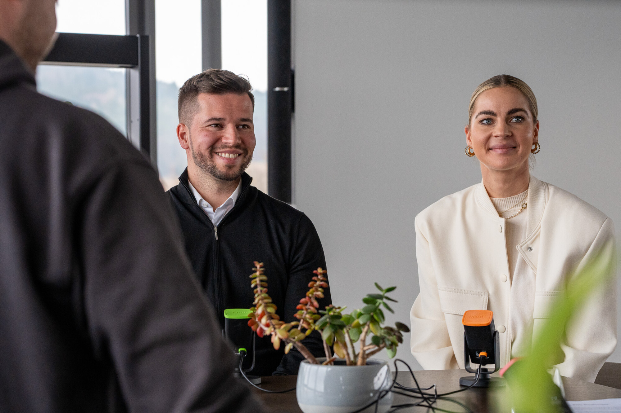 Zwei lächelnde Personen beim Podcast-Interview mit Mikrofonen vor einer Fensterfront und grünen Pflanzen.