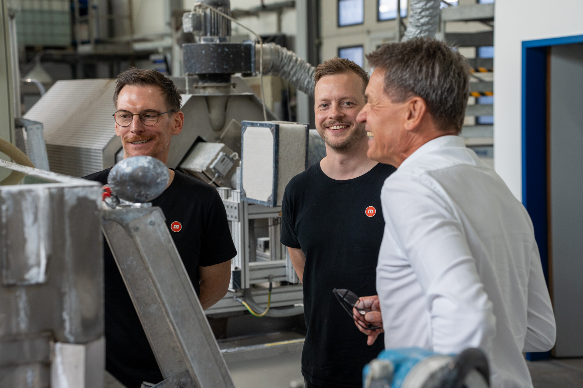 Drei Männer in Industriehalle im Gespräch, zwei tragen medienreaktor-T-Shirts, entspannte und positive Stimmung.