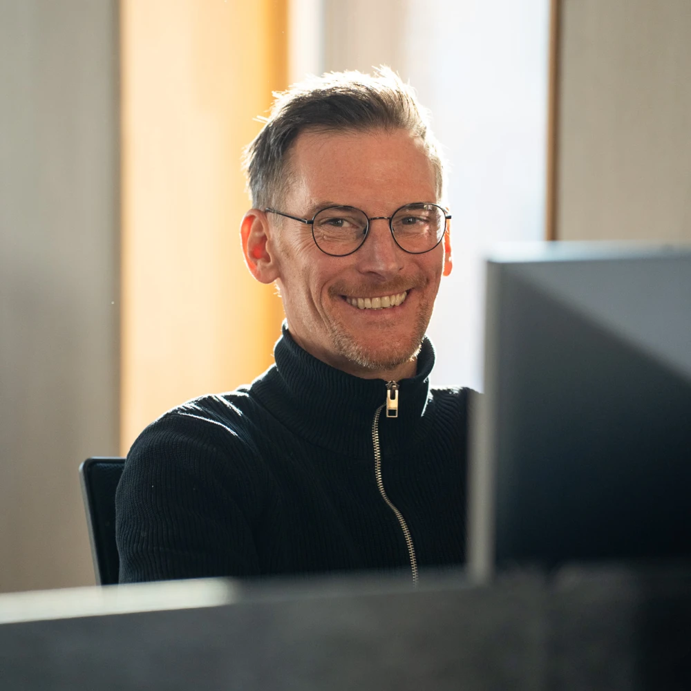 Mann mit Brille und schwarzem Rollkragenpullover lächelt am Arbeitsplatz vor Computerbildschirm in hellem Büro.