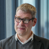 Mann mit Brille in Business-Kleidung lächelt freundlich vor hellem Bürofenster, professionelle Porträtaufnahme.