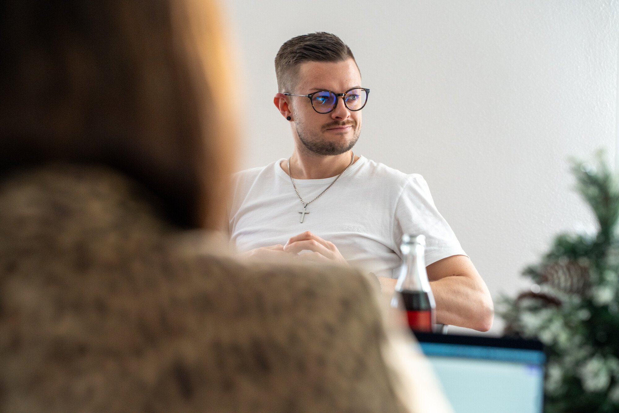 Mann mit Brille und weißem T-Shirt sitzt nachdenklich in modernem Büro, Kollege im Vordergrund unscharf.
