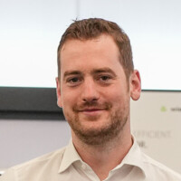 Junger Mann mit kurzen braunen Haaren und Bart lächelt freundlich, trägt weißes Hemd vor Bürohintergrund.