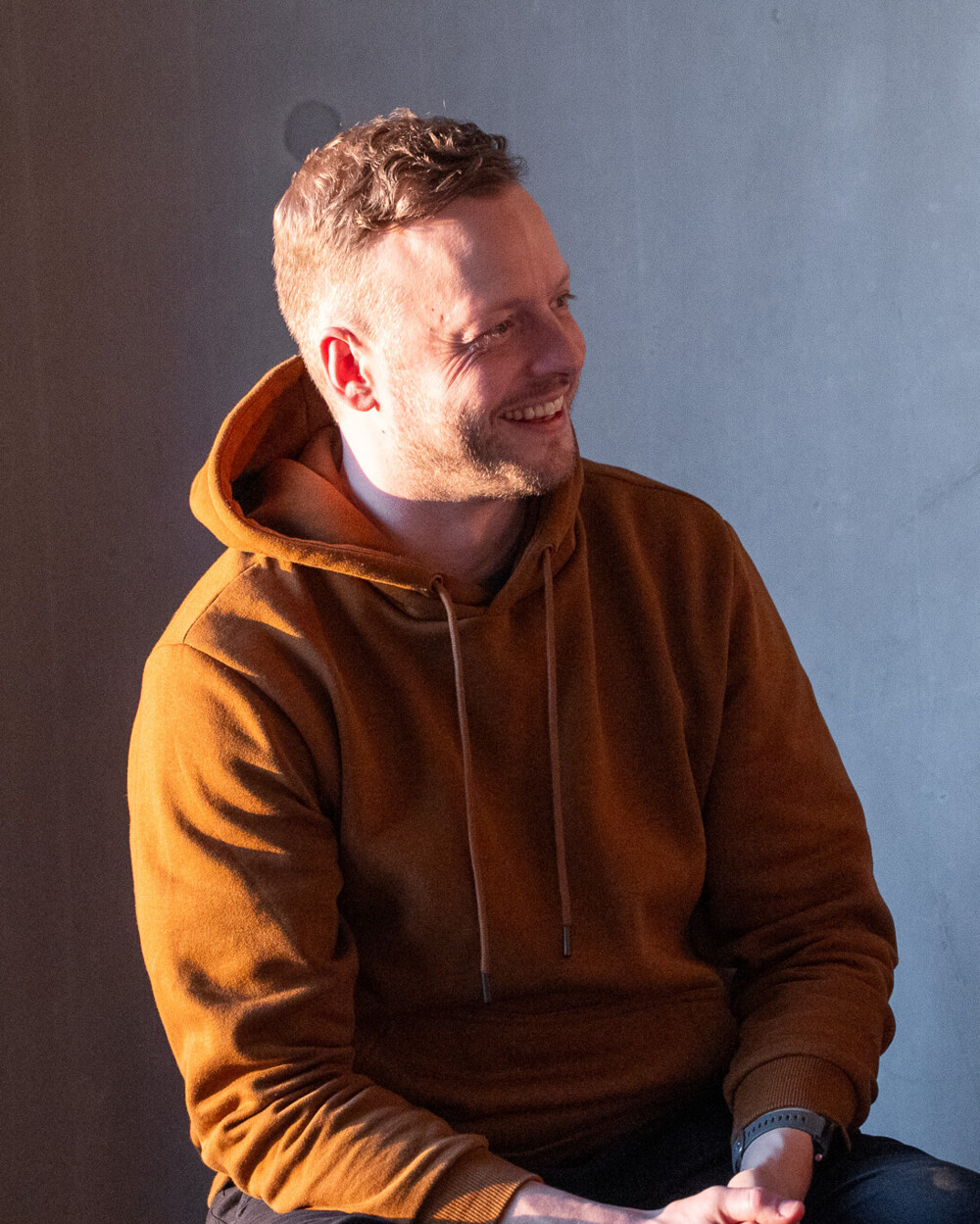 Lächelnder Mann im hellbraunen Hoodie sitzt entspannt vor grauem Hintergrund, natürliche Lichtstimmung betont Gesicht.