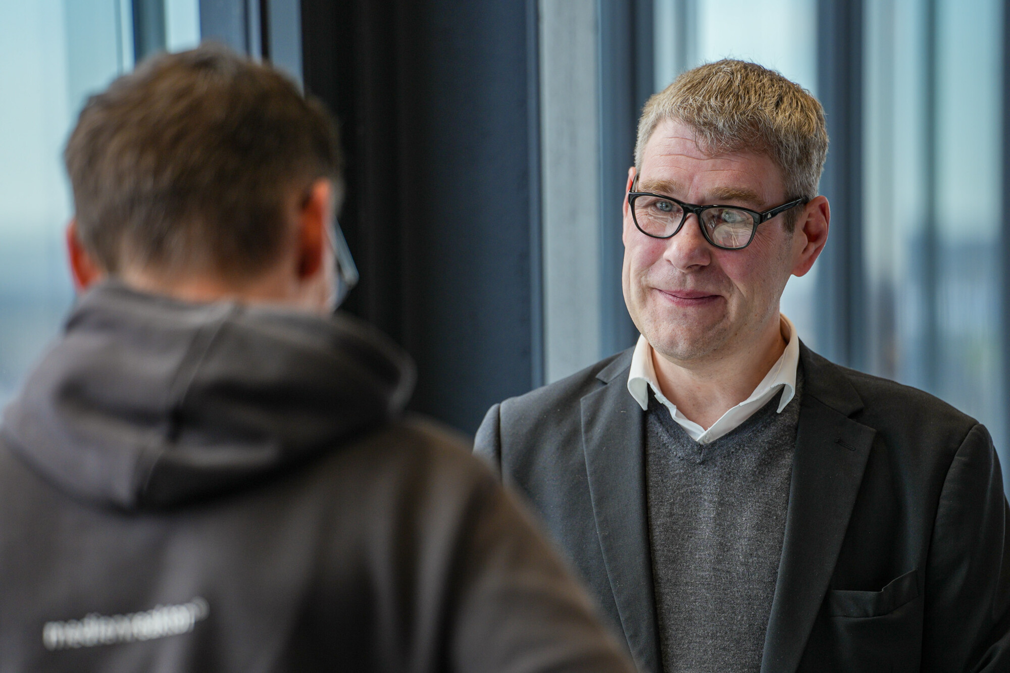 Mann im Anzug mit Brille im Gespräch mit Person in Hoodie, moderne Büroatmosphäre, freundliche Interaktion.