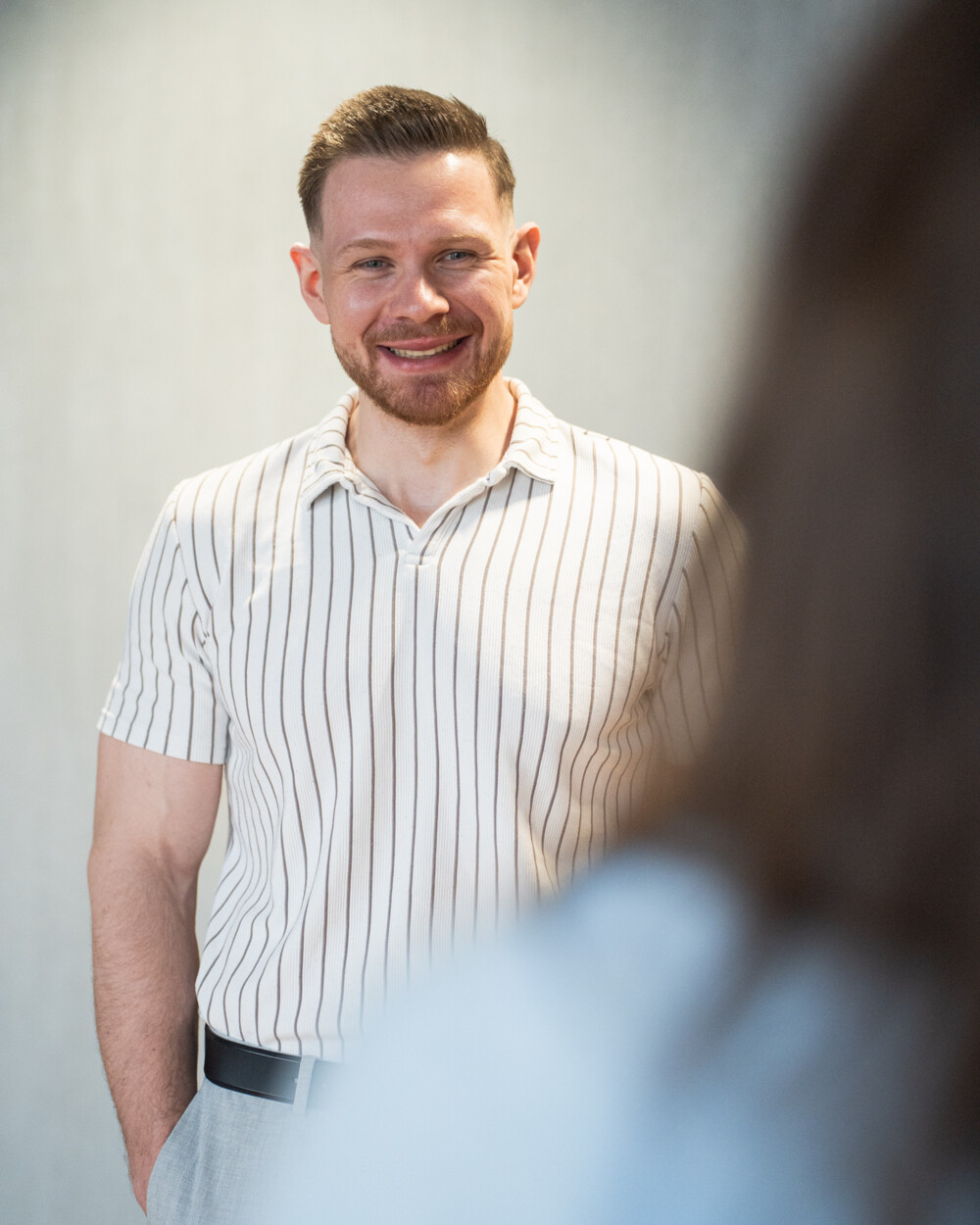 Junger Mann im gestreiften Poloshirt lächelt freundlich in Gesprächssituation, helle Bühne, positive Atmosphäre.