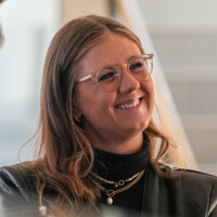Junge Frau mit Brille und Kette lächelt freundlich, casual Business-Umgebung, natürliches Licht, positive Stimmung.