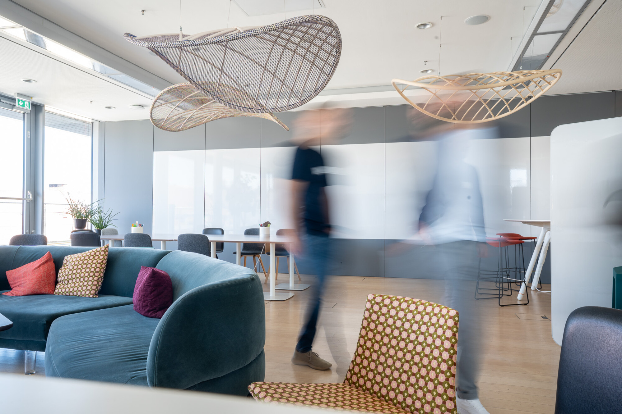 Moderne Lounge mit blauen Sofas, stilvollen Holzleuchten und bewegten Personen im hellen Büroraum.
