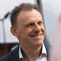 Mann mittleren Alters mit offenem Lächeln und lässigem Sakko in hellem Outdoor-Umfeld, sympathisch und freundlich.