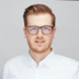 Junger Mann mit braunem Haar und Brille in weißem Hemd vor hellem Hintergrund, freundlich und professionell.