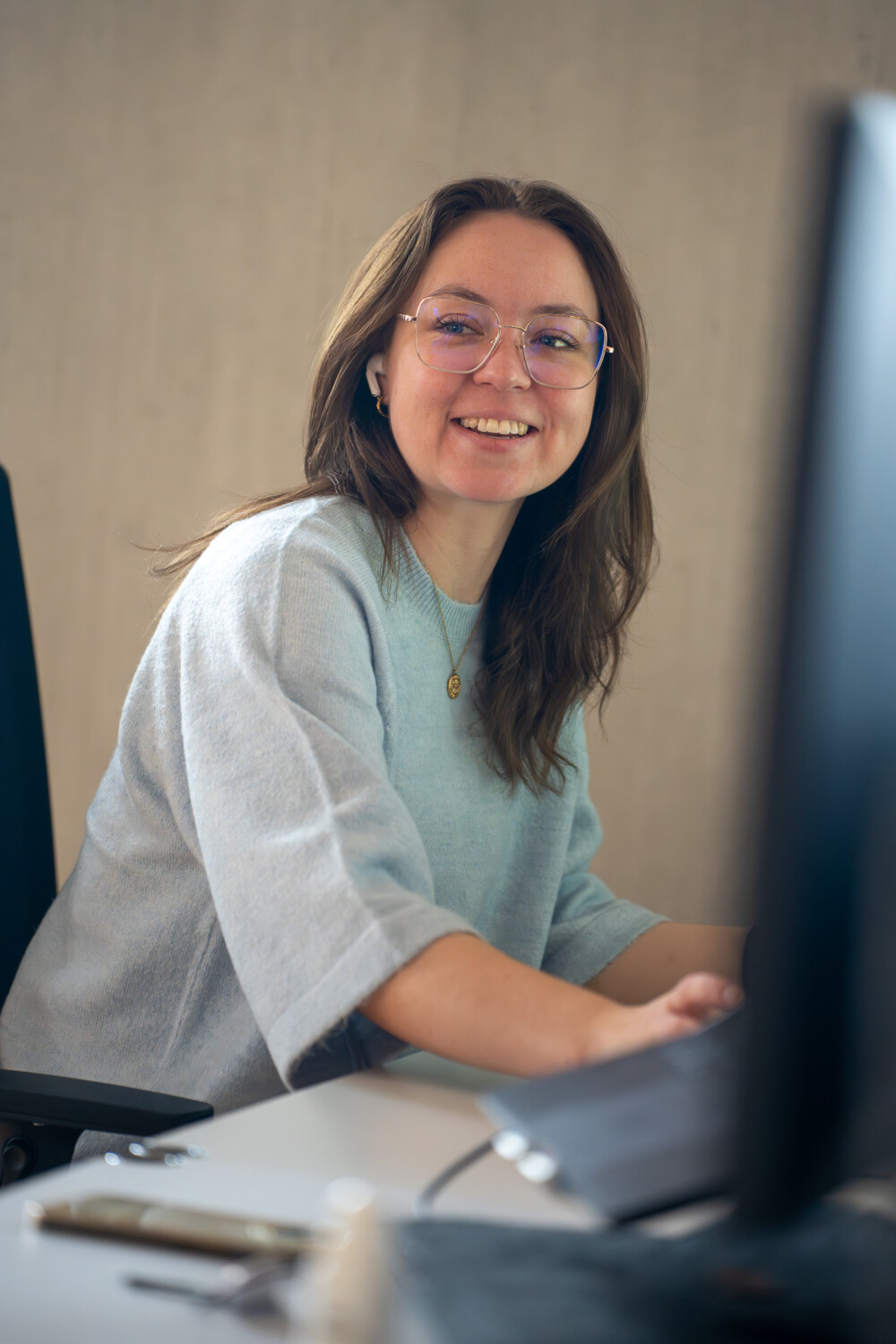 Junge Frau mit Brille und grauem Pullover arbeitet lächelnd am Laptop im modernen Büro.