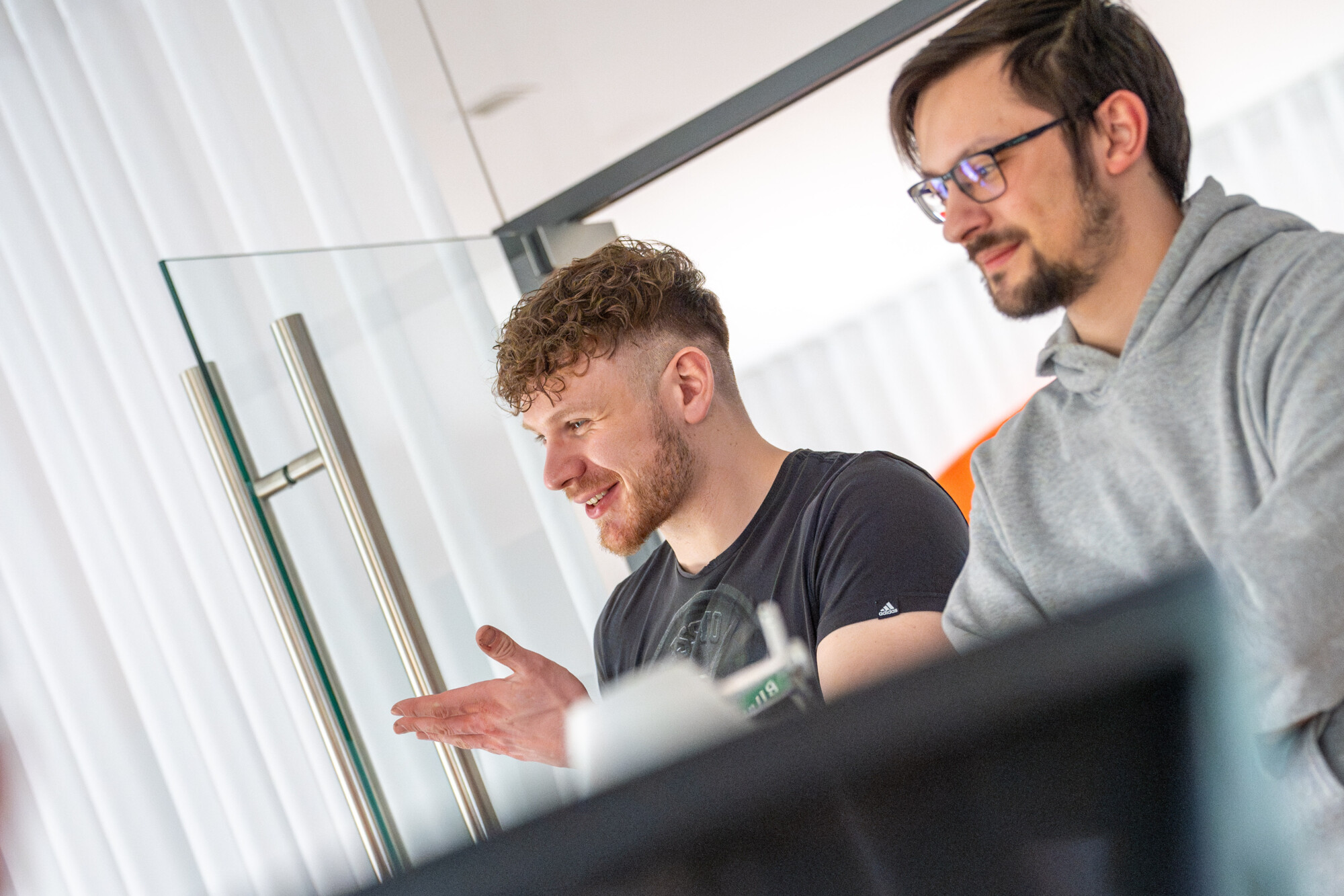 Zwei junge Männer im Büro diskutieren konzentriert und lächelnd an einem modernen Arbeitsplatz.