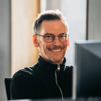 Fröhlicher Mann mit Brille und kurzem Haar in schwarzem Pullover vor hellem Hintergrund lächelnd.
