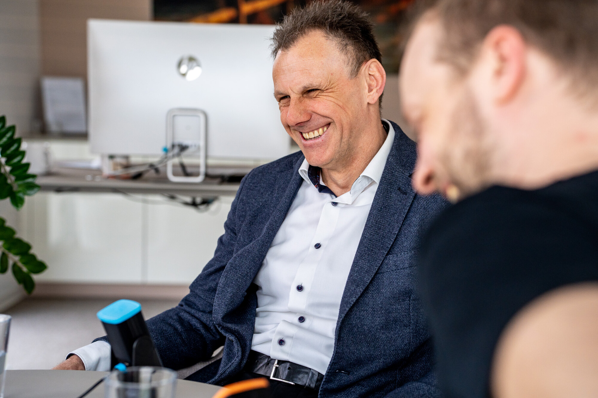 Geschäftsmann in blauem Sakko lacht herzlich während Meeting in modernem Büro mit Kollegen.